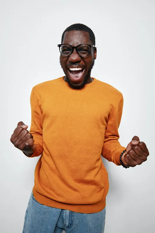 African American man with dark skin wearing glasses and orange sweater celebrating success with clenched fists and joyful expression on white background. Casual style and happiness.