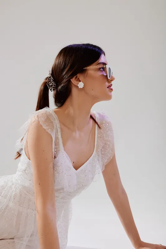 Young woman in a white lace dress wearing glasses poses in profile on a light background. Concept of beauty, fashion, elegance, and modern style with soft natural lighting.