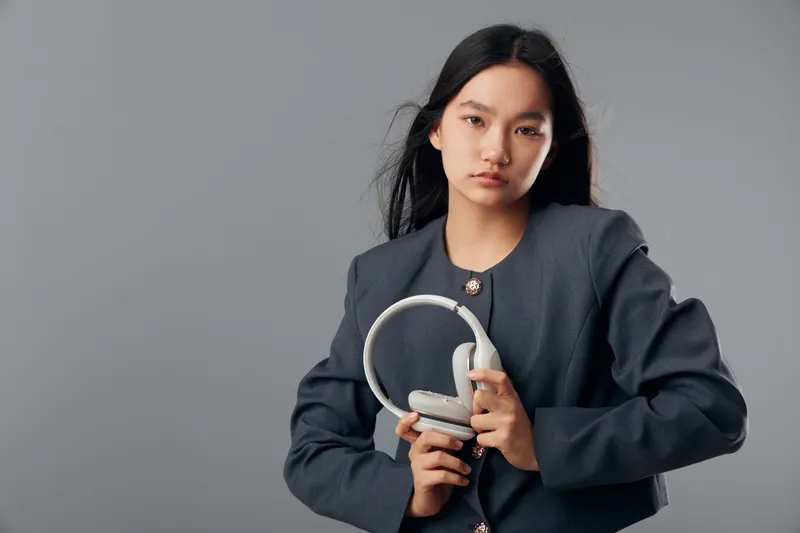Young woman in gray jacket holding headphones in hand posing on gray background. Serious female model with long black hair wearing casual outerwear in studio with copy space.