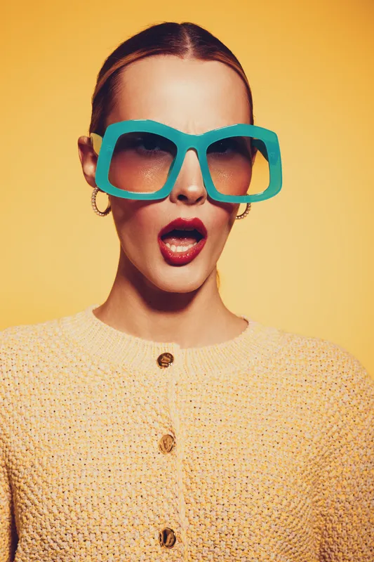 Surprised woman wearing large turquoise sunglasses and yellow knitted sweater poses against yellow background. Concept of fashion, style, summer accessories, and trendy eyewear.