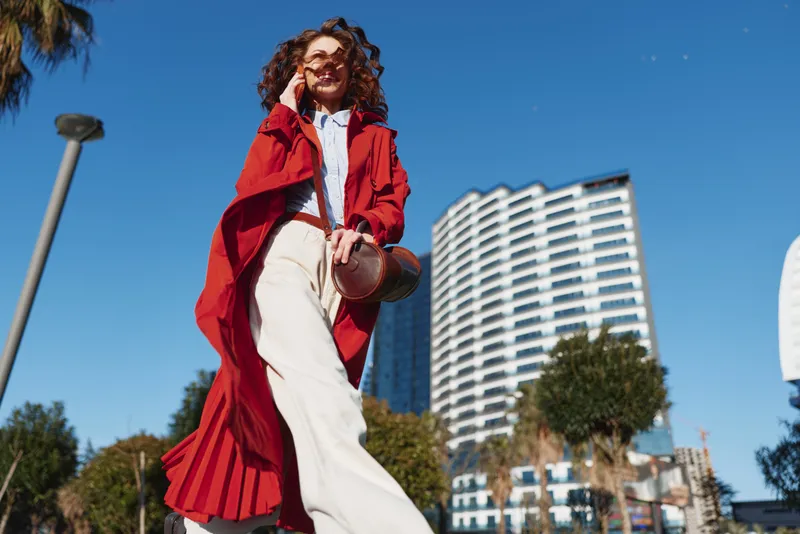 Young woman with curly hair strides through a sunny city street, wearing a red trench coat over white wide-leg pants, smiling as the camera captures her dynamic motion, evoking bold street style and