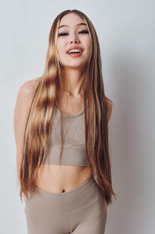 Young woman with long hair wearing beige sportswear, smiling and standing against white background. Fitness apparel, beauty and health concept with casual pose and natural light.