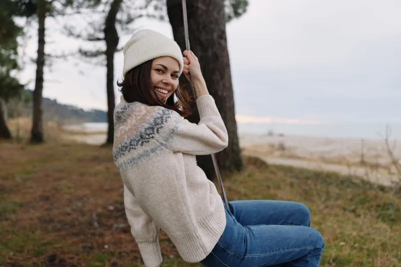 Woman on a swing in a cozy outdoor setting, smiling as she enjoys a casual moment by trees and a lakeside view, wearing a knit sweater and beanie for autumn comfort.