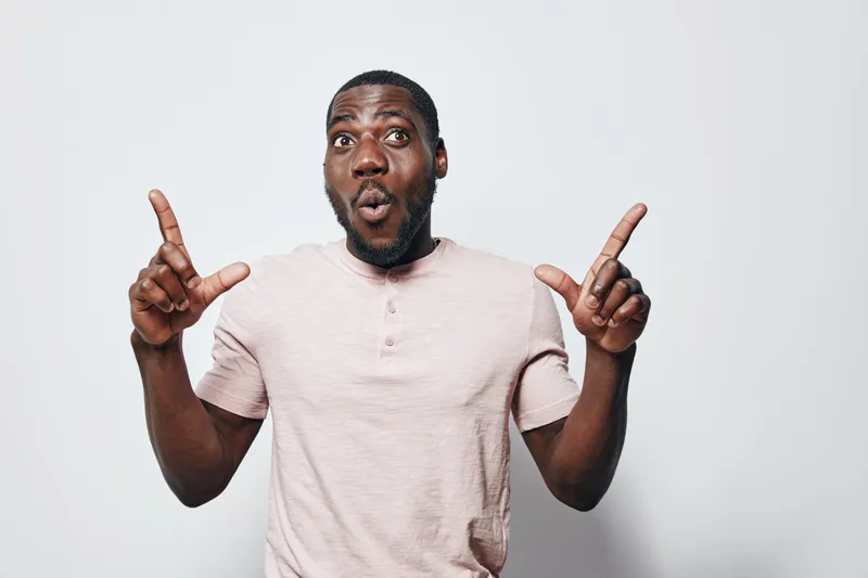 African American man with dark skin wearing light pink shirt showing surprised expression with raised eyebrows and pointing fingers up on white background. Male adult, casual style.