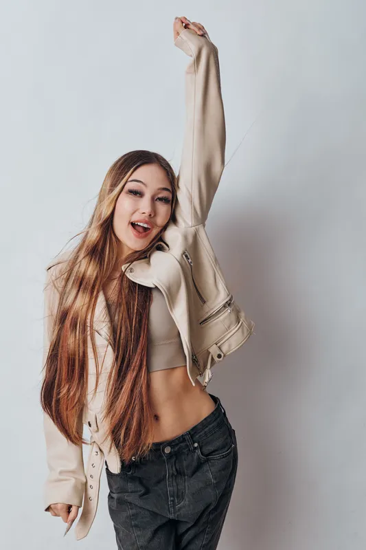 Young woman with long hair wearing casual jacket and crop top raising arm and smiling in studio. Fashionable girl posing with cheerful expression on gray background for lifestyle portrait.