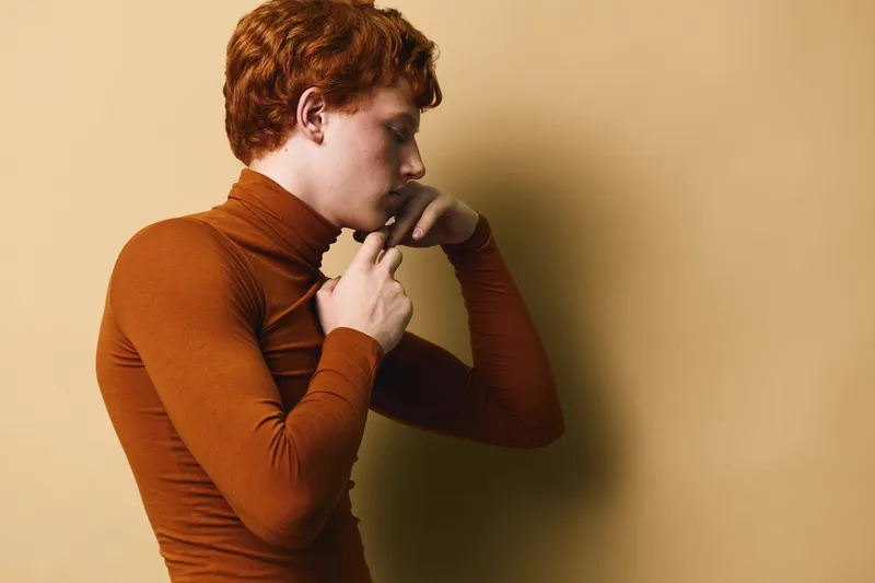 Young man with red hair wearing brown turtleneck, posing with hand near face, looking thoughtful on beige background in studio lighting.