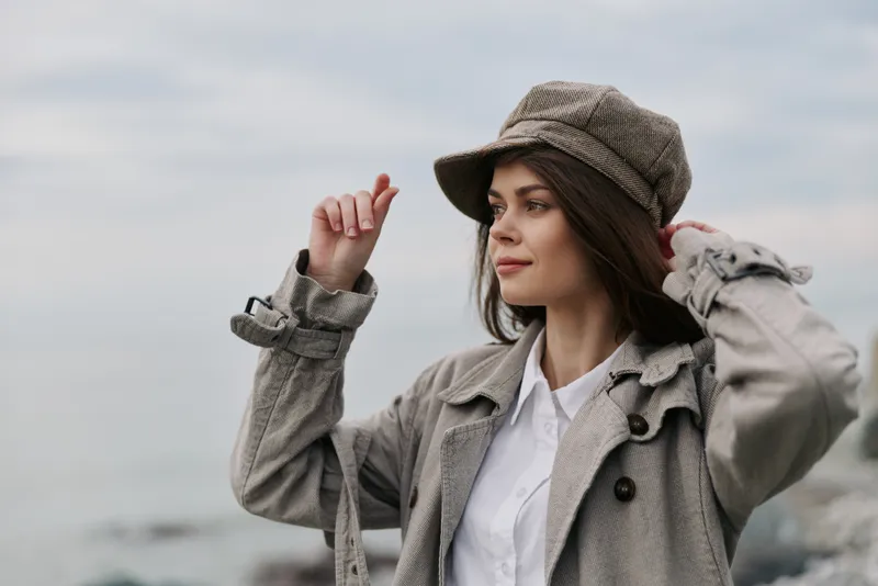 Young woman in a beige trench coat and checkered hat adjusts her hat outdoors near a calm sea under a cloudy sky. Casual autumn fashion and nature background concept.