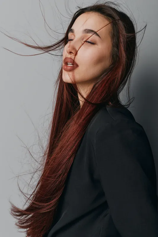 Woman with long red hair and closed eyes posing in black jacket on gray background. Portrait of female model with natural makeup and wind blowing hair in studio setting.