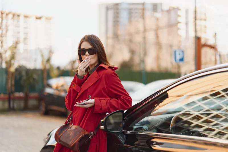 Stylish woman in a red coat stands beside a black car, covering her mouth with her hand, in an urban setting with tall buildings and parked cars.