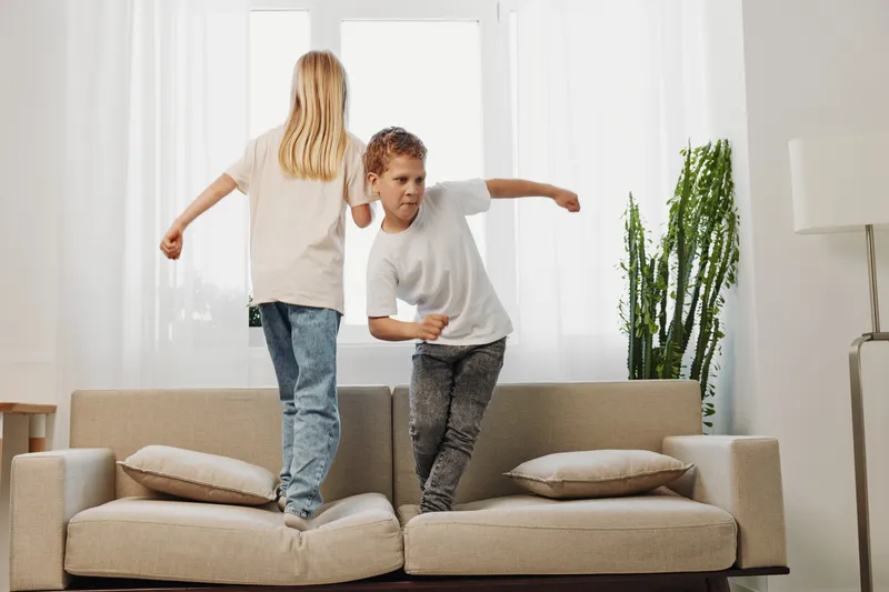 Two children jumping on a sofa in a bright living room, having fun and playing actively. Casual home scene with natural light and modern furniture.