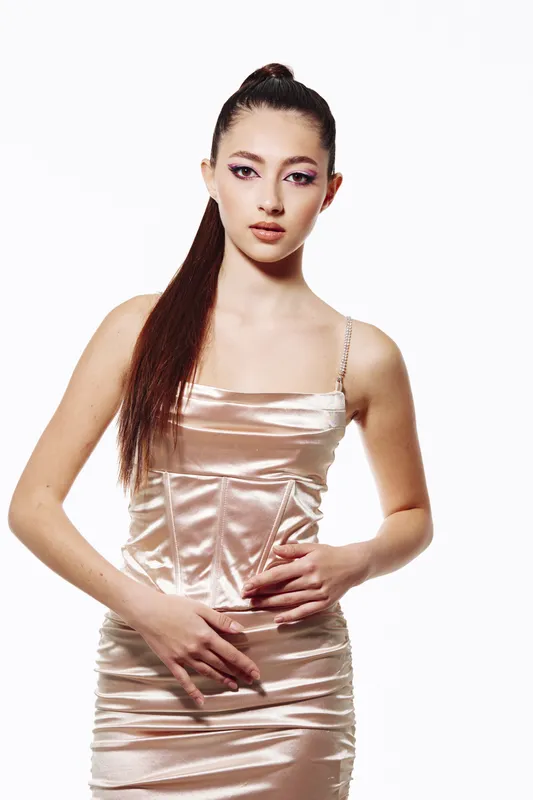 Young woman in a shiny satin dress poses confidently on a white background with sleek high ponytail and elegant makeup. Creativity and fashion concept focused on beauty trends.