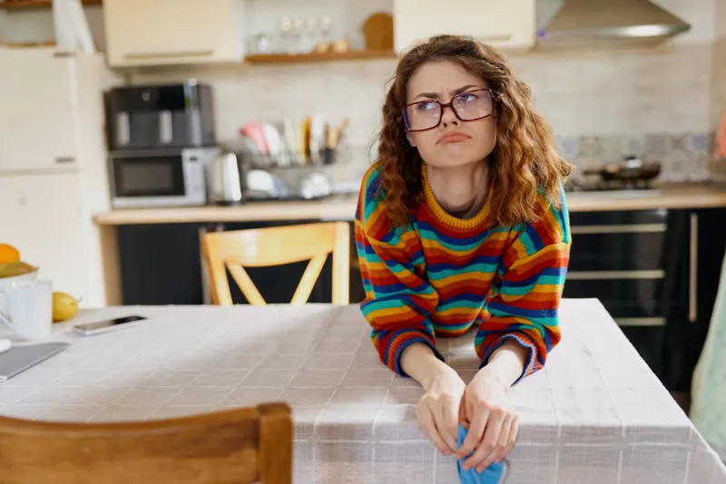 Woman in a colorful striped sweater sits at a kitchen table with glasses, a contemplative expression, capturing a home lifestyle moment in a cozy domestic setting.