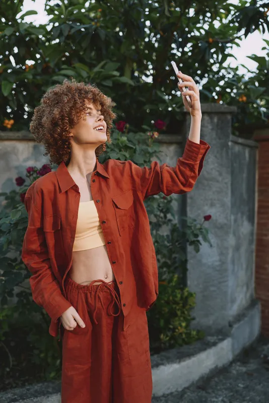 Cheerful woman outdoors selfie fashion casual confident rust outfit sunny nature greenery pose expression smile breeze street urban backdrop sunlight portrait stylish relaxed colorful vibrant earth