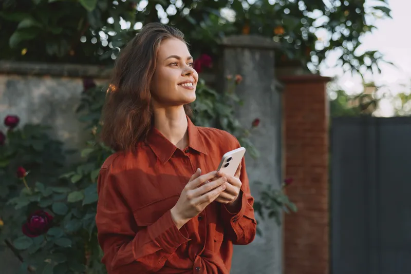 Woman lifestyle outdoor portrait smiling, she holds smartphone and wears a rust shirt, relaxed mood, garden backdrop with greenery, candid moment of casual fashion and everyday communication