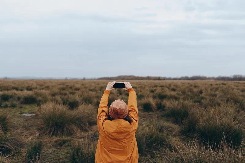 Woman outdoors lifestyle in meadow field, orange coat and knit hat, hands adjusting hat near head, nature calm, soft cloudy light, tall grass background