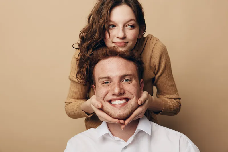 Smiling young couple with woman holding man’s face gently. Warm beige background, casual clothing. Concept of love, affection, happiness, and close relationship between partners.