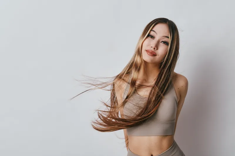 Young woman with long hair wearing a beige crop top and leggings posing against a plain light background. Fashion model with natural makeup and casual sporty outfit in studio.