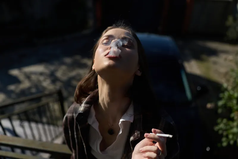 Young woman outdoors smoking a cigarette, wearing a casual jacket, head tilted back, relaxed urban mood, candid lifestyle portrait with natural light, sunlit environment