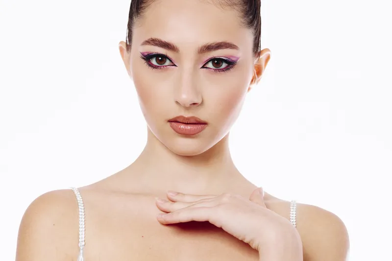 Beautiful young woman with creative purple eye makeup and glossed lips poses elegantly with hand near chin on white background. Concept of beauty creative fashion and makeup artistry.