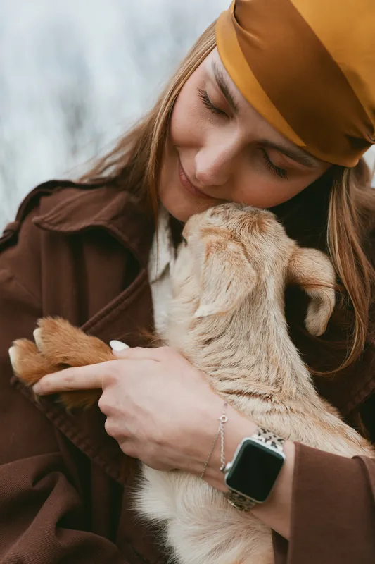 Warm lifestyle portrait of a woman in a brown coat and headband cuddling a goat kid outdoors, showing tender affection, rustic charm, and a cozy mood under soft natural light