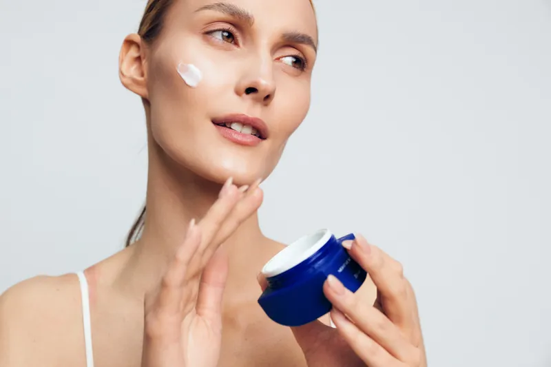 Young woman applies face cream on cheek holds blue jar skincare beauty routine isolated on white background natural healthy skin care concept closeup portrait.