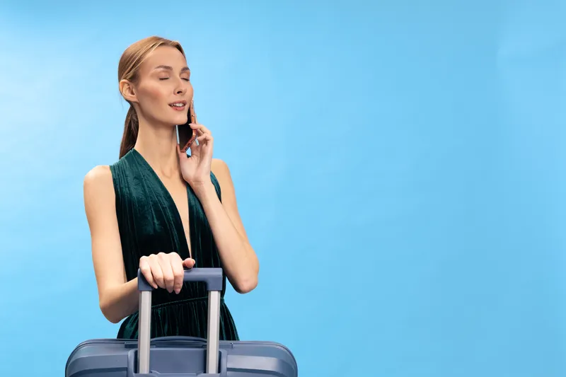 Young woman in green dress holding suitcase and talking on phone with closed eyes on blue background. Concept of travel, communication, and vacation planning.