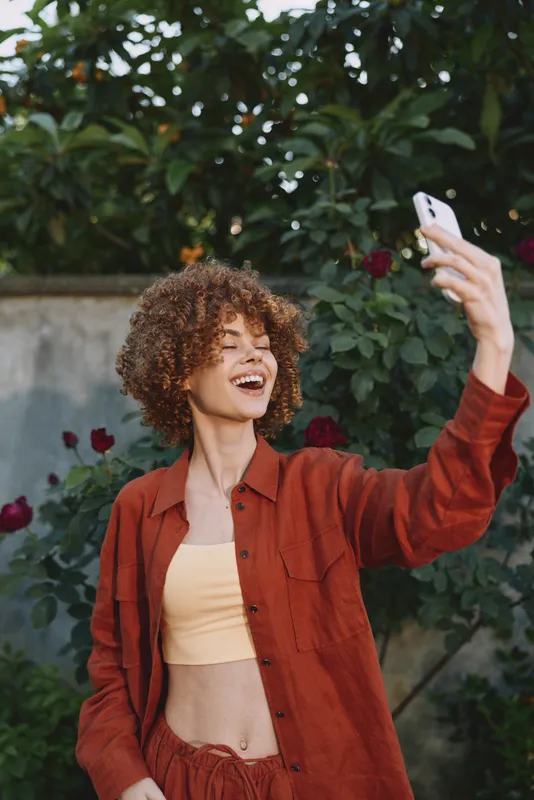 Smiling woman taking a selfie in an outdoor setting, wearing a rust jacket over a beige crop top. Lifestyle and fashion shot with cheerful self-expression in a lush garden