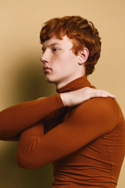 Redhead man in brown turtleneck sweater looking sideways with arms crossed on chest in calm thoughtful pose against beige background. Profile portrait of young male in casual outfit.