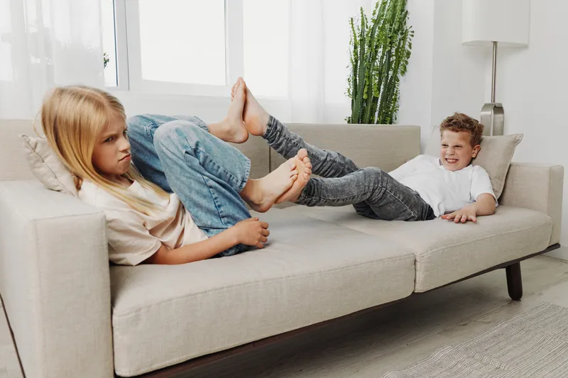 Two children lying on a sofa, playfully interacting with their feet, showing sibling rivalry and playful mood in a bright living room.