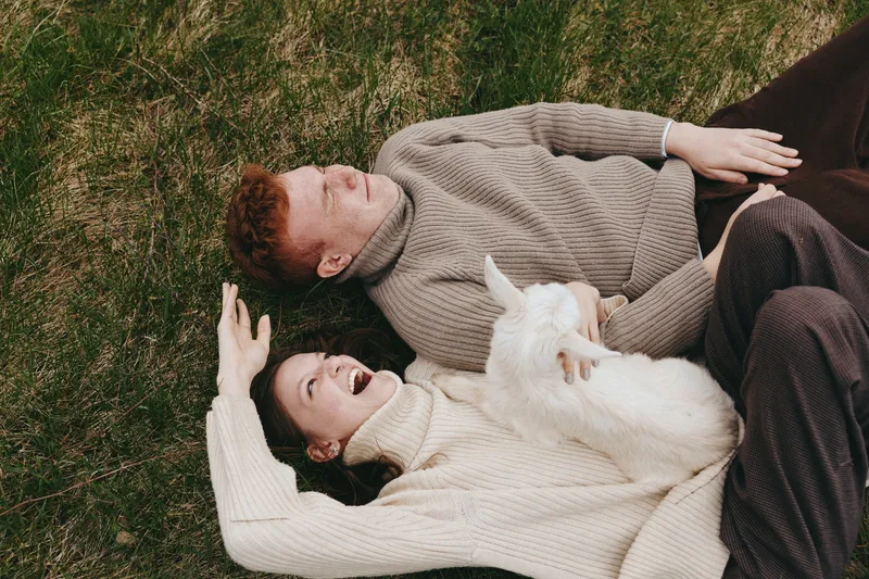 Smiling couple lying on grass outdoors with a fluffy white pet, enjoying relaxed moments together. Cozy sweaters, natural light, calm mood, friendship and wellness themes. Close, intimate scene
