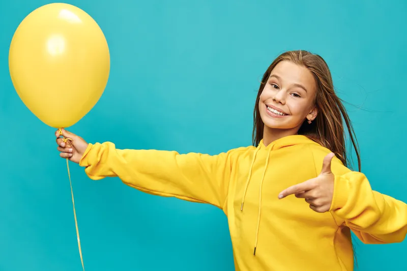 Girl with yellow hoodie holding yellow balloon pointing with finger on bright turquoise background smiling cheerful confident casual clothing joyful child celebration.