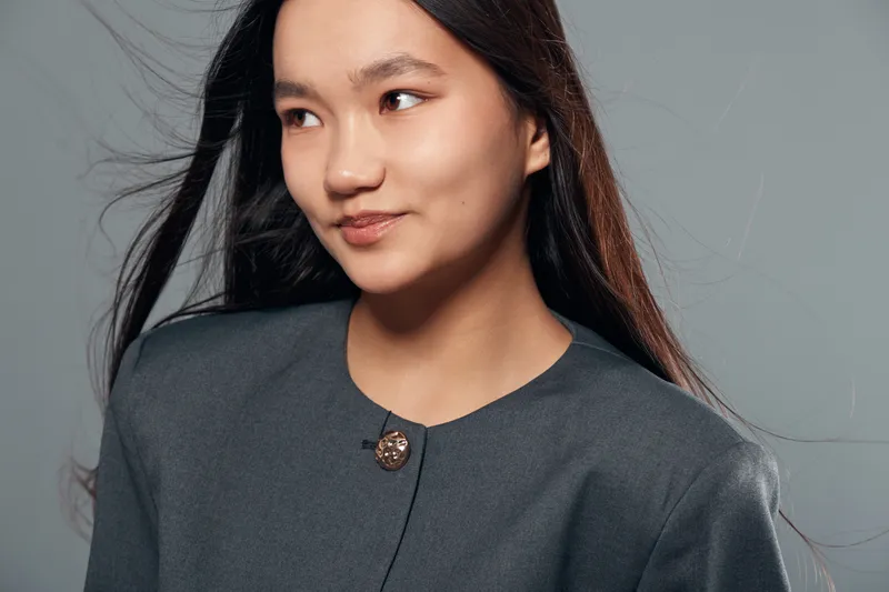 Portrait of young Asian woman with long hair wearing grey jacket looking sideways against neutral background. Female model with natural makeup and subtle smile in studio lighting.