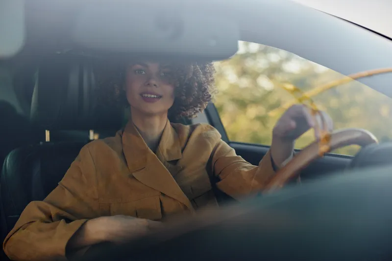 A woman drives a car in warm retro film tones, capturing a lifestyle moment. The interior glow and confident smile convey independence and everyday travel, candid and stylish.