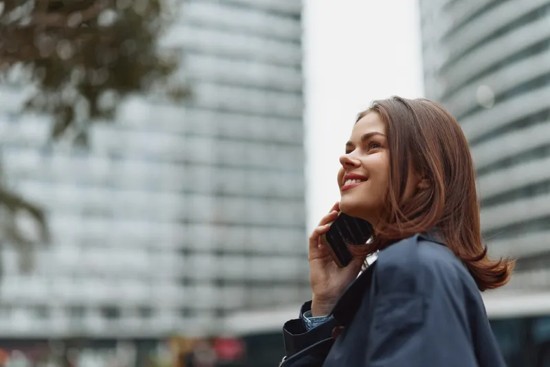 Young woman in a navy blazer chats on a smartphone in a bustling city, smiling as tall glass buildings blur in the background, appearing confident and engaged.