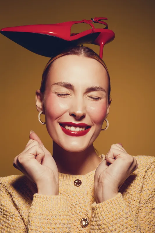 Happy woman with closed eyes and red lipstick smiles joyfully while balancing a red high heel shoe on her head. She wears a yellow sweater and gold hoop earrings. Concept of fun and fashion.