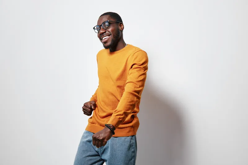 African American man with dark skin wearing glasses and orange sweater dancing happily against white background. Casual style, smiling and enjoying music or celebration.