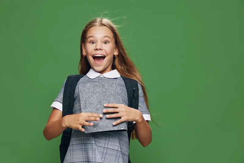 Girl with long hair in school uniform holding a book and wearing backpack smiling with excitement on green background for education, learning and childhood concept.