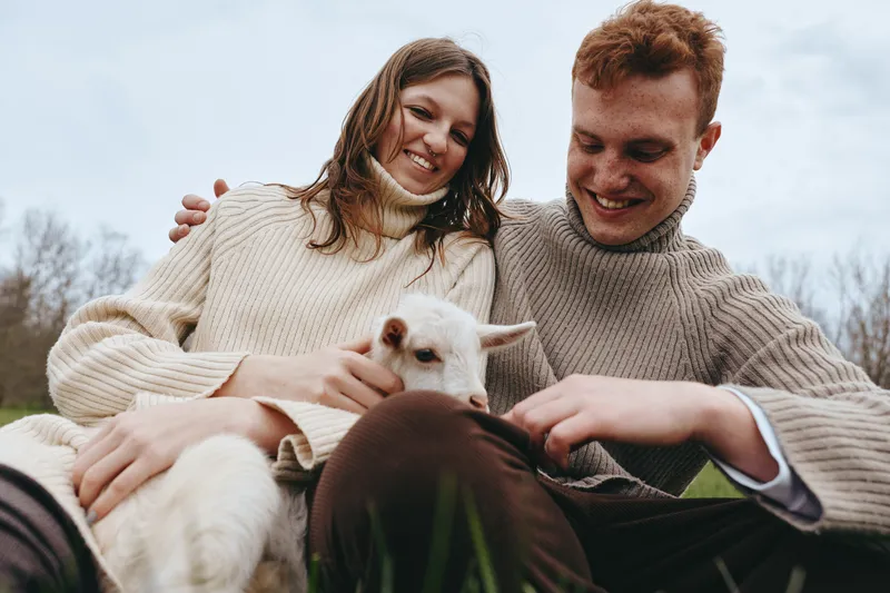 A couple enjoys a playful moment with a friendly goat kid outdoors, wearing cozy sweaters, smiling warmly, capturing companionship and rustic lifestyle in a sunny countryside scene.