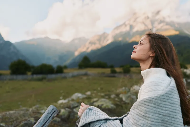 Young woman with long hair wrapped in a white knit sweater enjoys a serene mountain landscape outdoors
