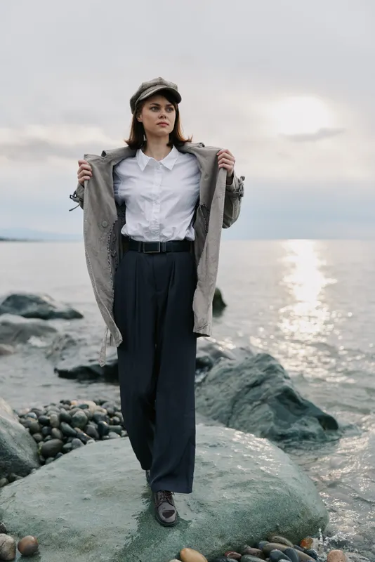 Woman in stylish casual clothes stands confidently on rocky shore near sea under cloudy sky. Fashion portrait with natural light and tranquil water background. Concept of autumn outfit.