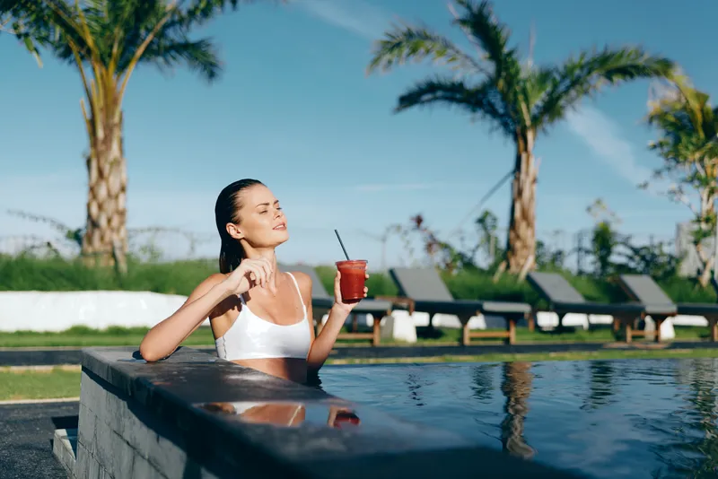 Relax pool scene with a woman in a white bikini savoring a cold beverage, enjoying sunny leisure by the water in a tropical outdoor setting, summer.