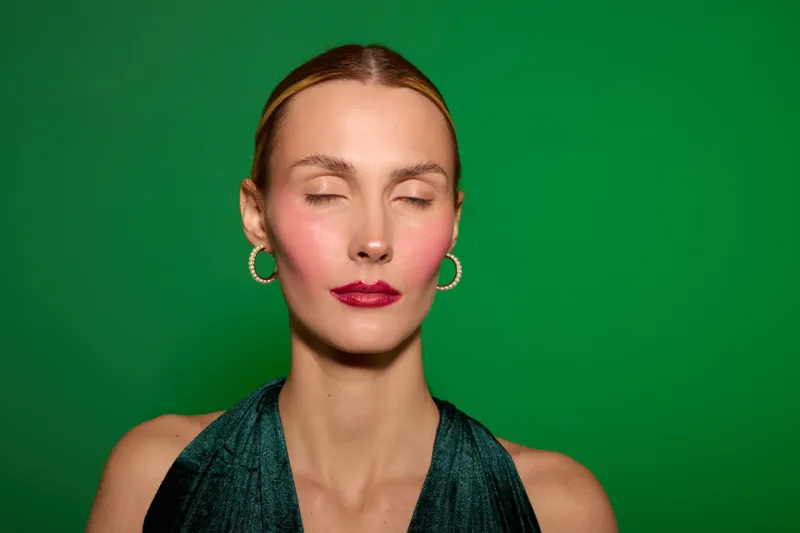 Young woman with closed eyes and red lipstick poses against green background wearing dark sleeveless top and gold hoop earrings. Concept of beauty, fashion, and calm expression.