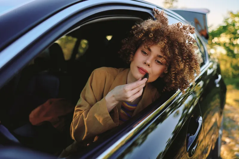 A woman leans from a car window to touch up makeup, casual chic style, warm retro film tones, capturing a candid moment of daily beauty routine and self care.
