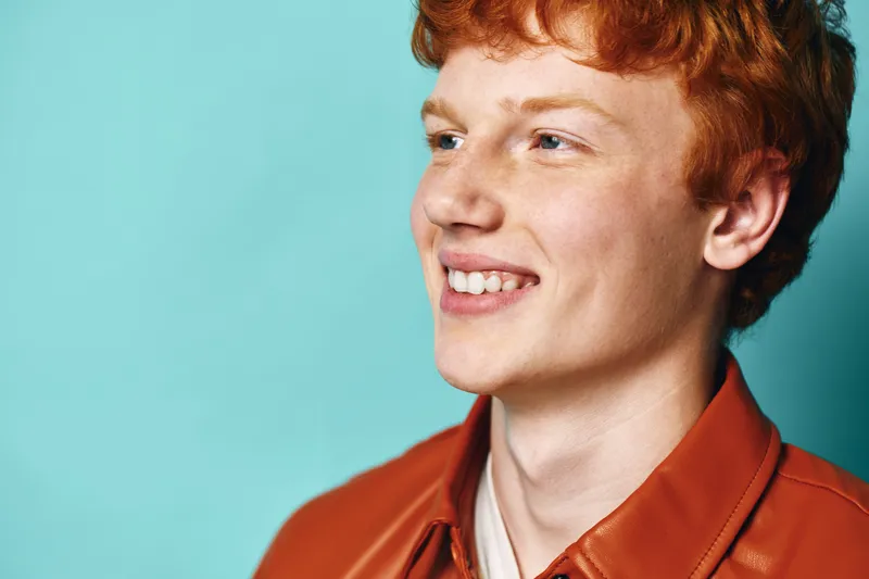Young man with red hair smiling and looking to side, wearing orange shirt against blue background. Portrait of cheerful male person with freckles and casual clothing in studio setting.