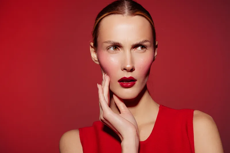 Young woman with red lipstick and blush on cheeks poses against red background wearing sleeveless top. Concept of fashion, beauty, makeup, and confident expression.