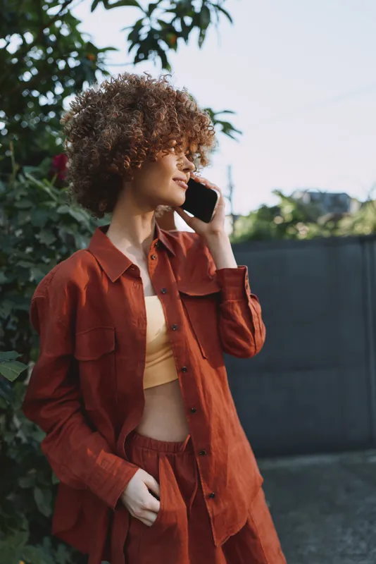 Casual woman with curly hair wearing orange outfit talks on phone outdoors in garden setting, capturing modern lifestyle and relaxed fashion mood for daily urban story, creative portrait