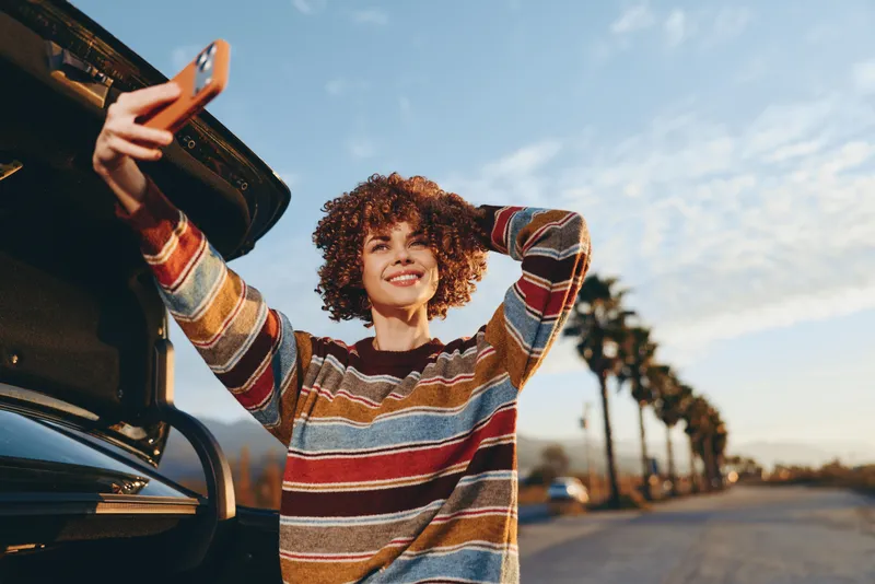 Woman lifestyle outdoors, smiling, curly hair, standing by a car at sunset, striped sweater with sleeves raised, relaxed mood, warm golden sunlight, palm road background, soft breeze