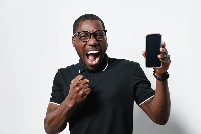 African American man with dark skin wearing glasses and black polo shirt showing smartphone with excitement and clenched fist on white background.