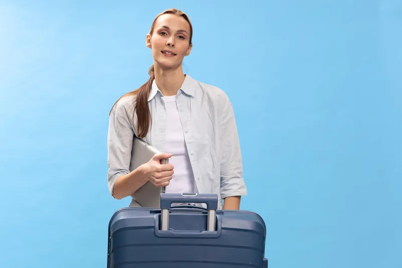 Young woman traveler with suitcase and laptop stands smiling on blue background. Concept of business trip, vacation, preparation, and travel planning with modern technology.
