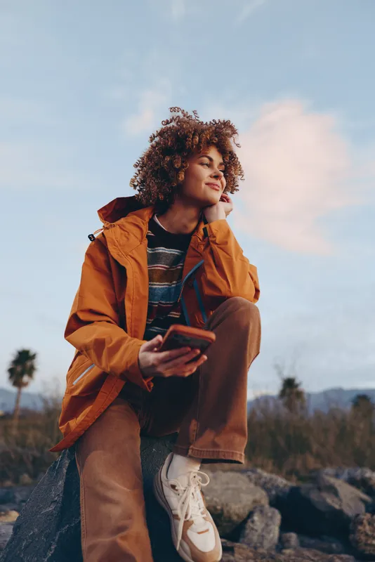 Woman lifestyle outdoors, curly hair, orange jacket, holding smartphone, smiling in warm sunset light, relaxed pose on rocky landscape, palm trees softly blurred background